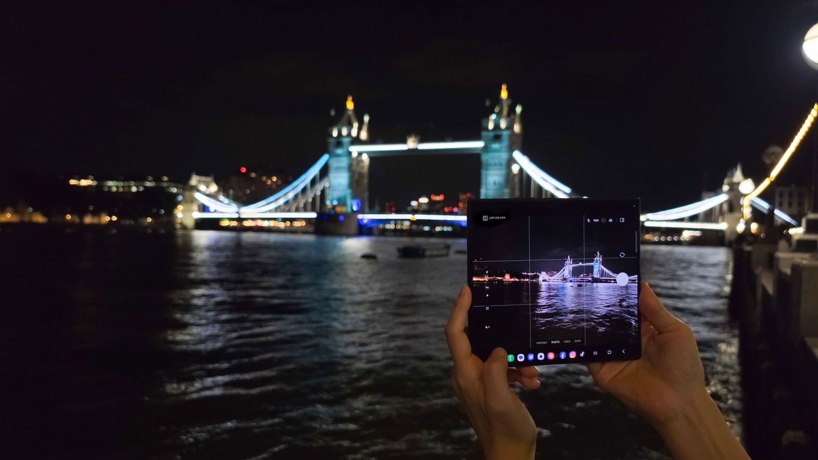 Ruce držící rozložený Samsung Galaxy Z Fold s otevřenou aplikací fotoaparátu, v pozadí noční Tower Bridge